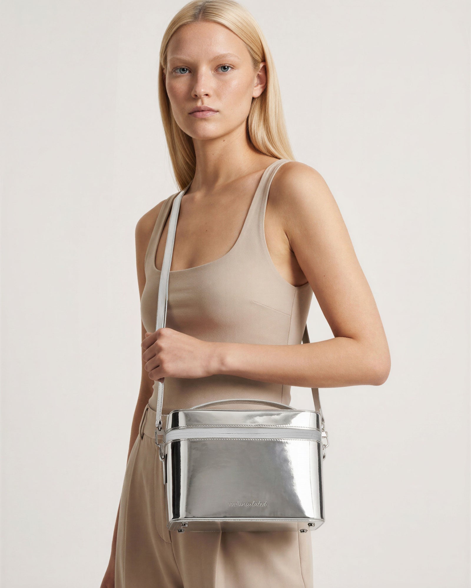 A blonde model  carrying a shiny silver metallic crossbody performance Beauty Case with strap.